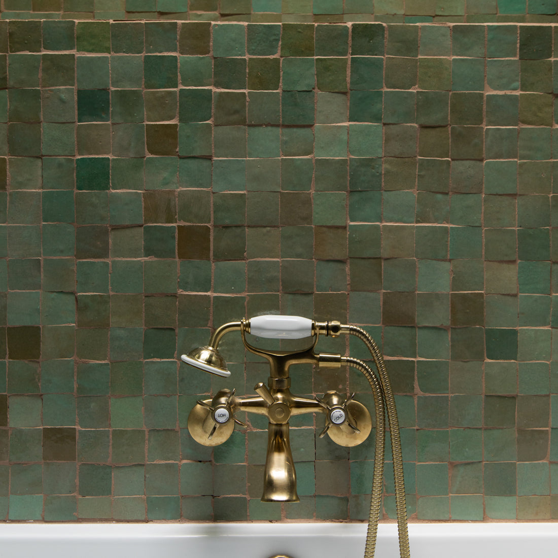 A brass bathtub faucet with white accents is mounted on a wall covered in green square tiles. The lower part of a white bathtub is visible at the bottom of the image.