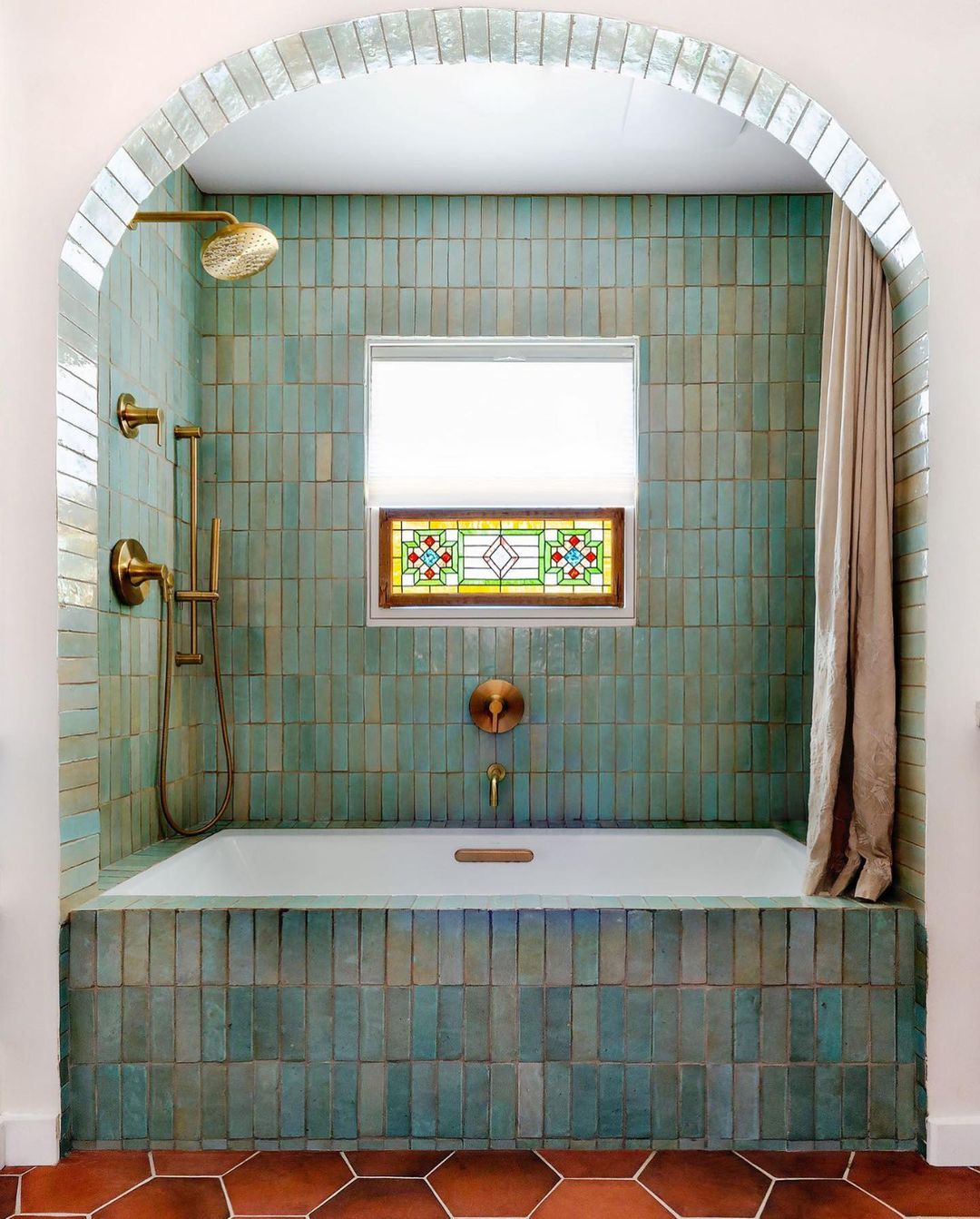 A bathtub with greenish-blue vertical tiles, brass fixtures, a window with a stained glass panel, and a white curtain, set within an arched alcove above terracotta floor tiles.