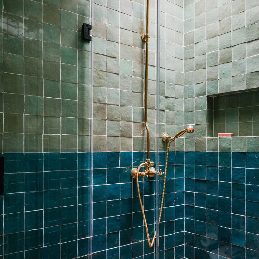 A shower with glossy green and teal square tiles, divided horizontally. The shower features a gold metal showerhead and fixtures, with a built-in shelf holding a red item in the wall above the darker tiles.