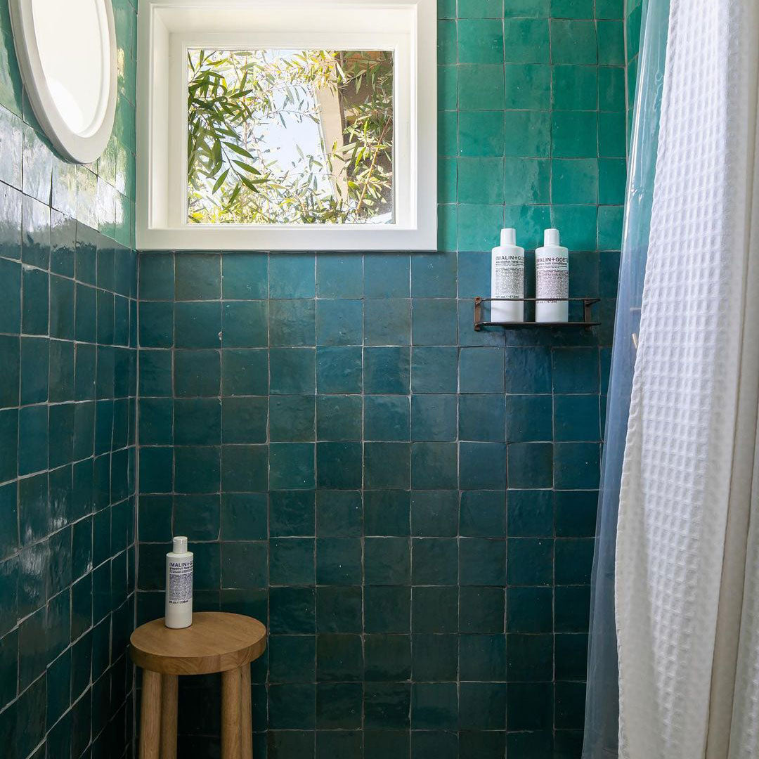 A shower with teal square tiles, a small window showing tree branches, a wooden stool with a bottle, a wall shelf with two bottles, and a white textured shower curtain partially visible on the right.