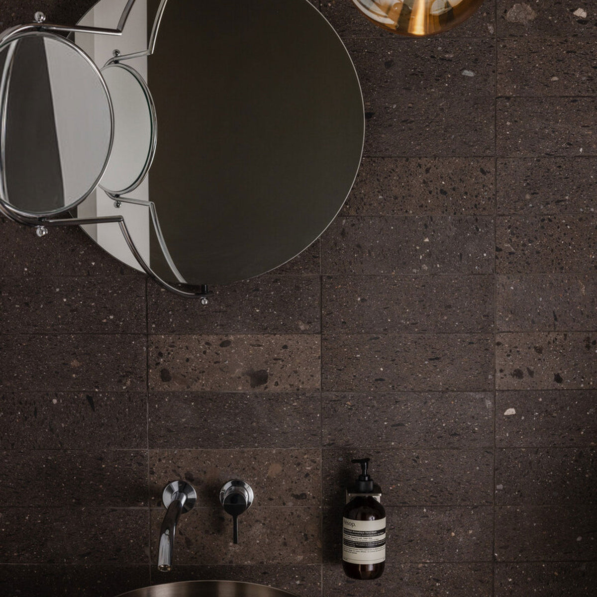 A round mirror and magnifying mirror hang above a dark stone bathroom sink, with a wall-mounted faucet and a bottle of liquid soap on the tiled wall.