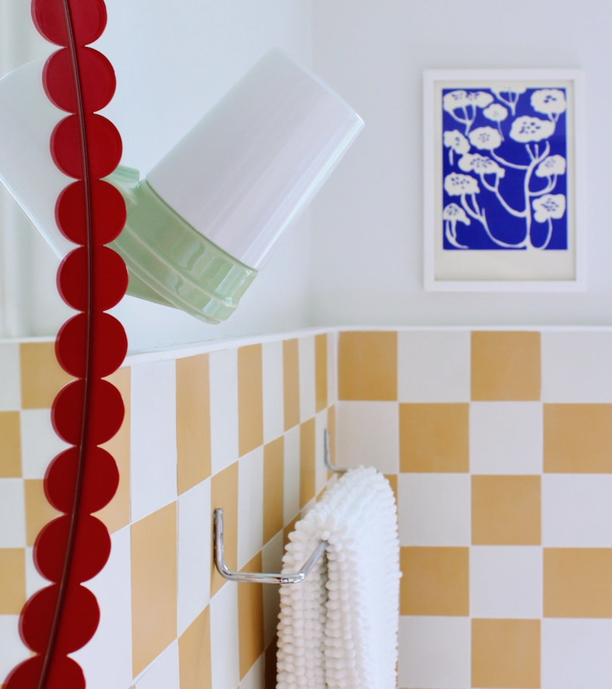 A bathroom features vendor-unknown's White 4x4 tiles in a yellow and white checkerboard pattern on the floor, plus a round red-edged mirror, white towel on metal rack, green-white lamp, and blue-and-white botanical print art.