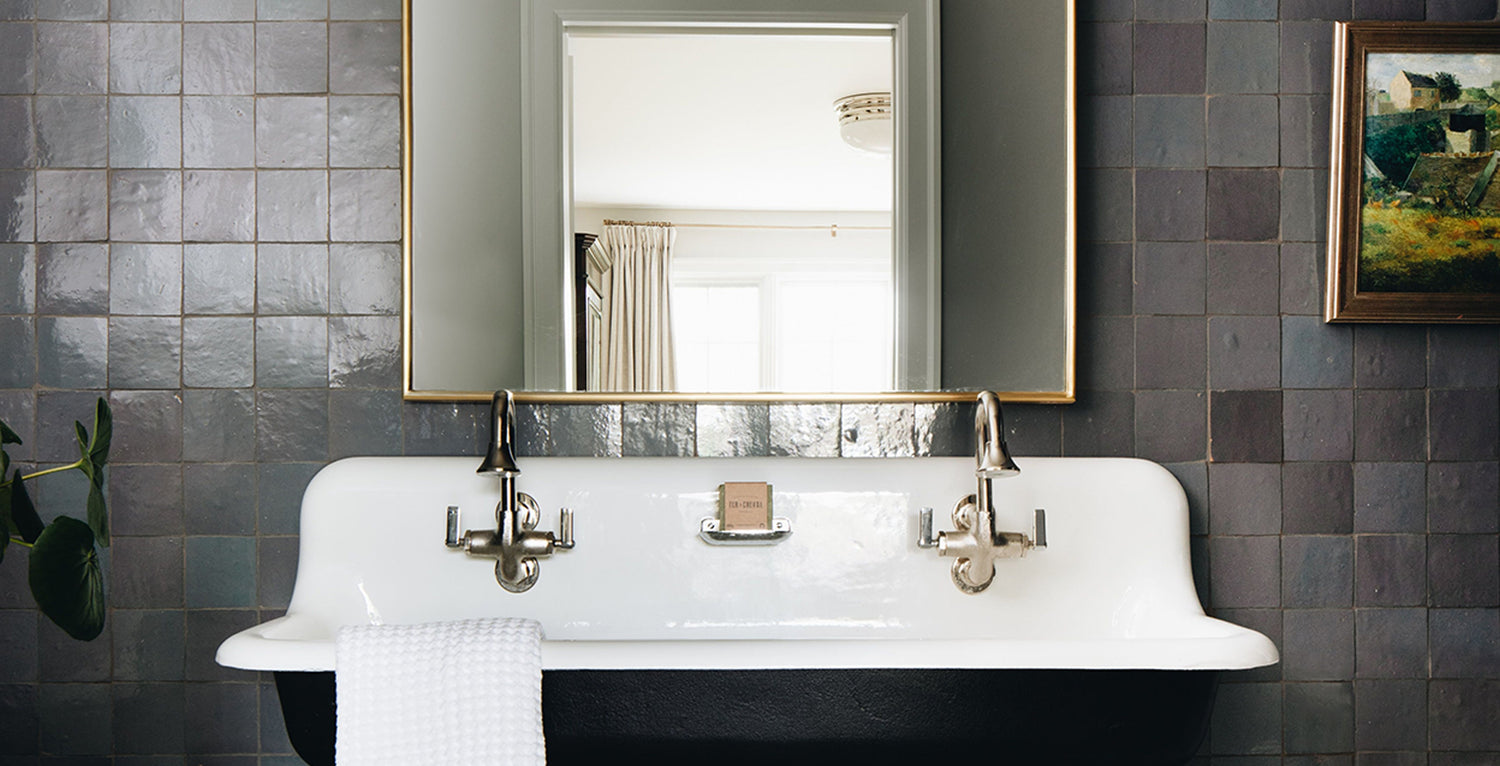 A vintage-style double sink with two faucets sits beneath a large mirror on a dark tiled wall. A white towel hangs on the edge, and a framed painting is visible on the right side of the image.