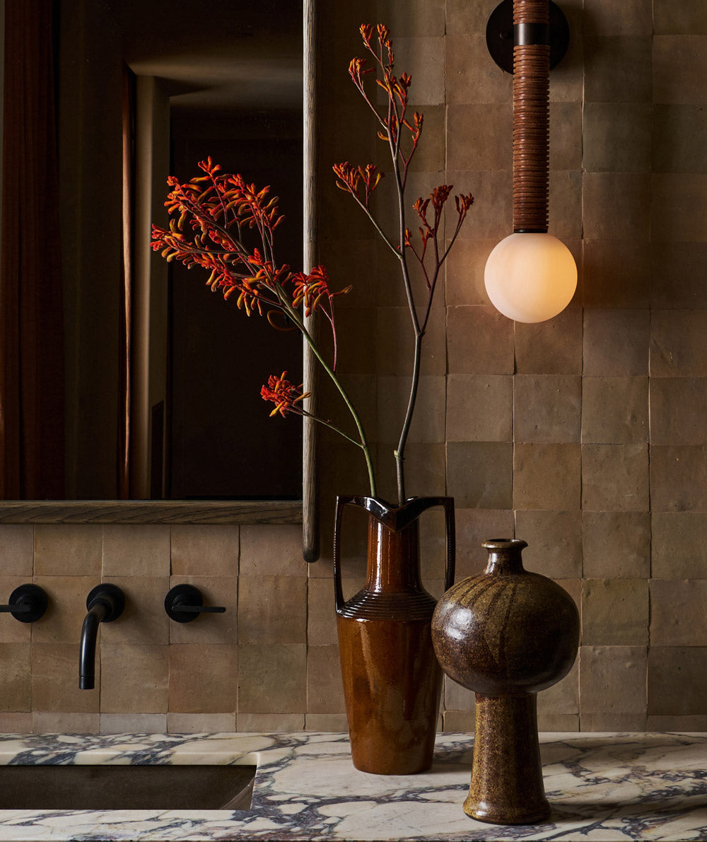 A modern bathroom with a marble countertop, brown ceramic vases with red flowers, a wall-mounted faucet, a large mirror, and a round wall sconce emitting warm light against textured brown tiles.
