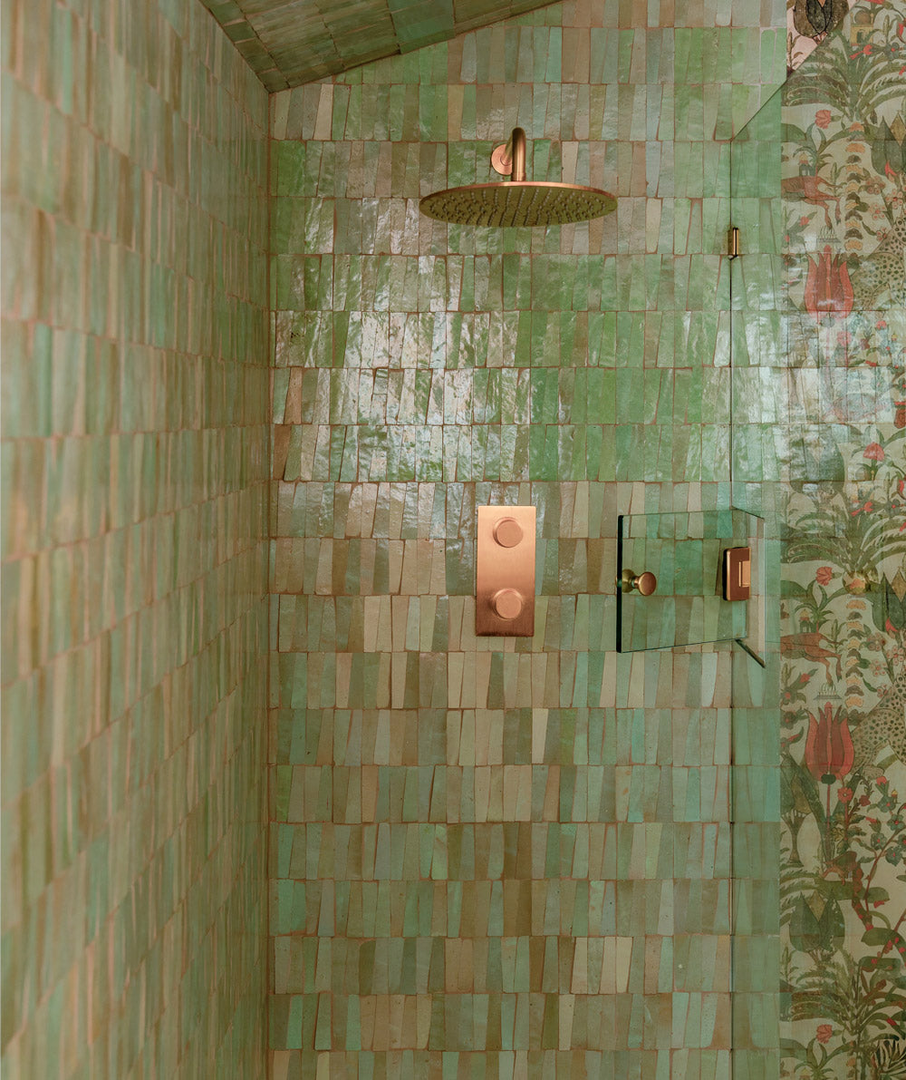 A shower with green, unevenly shaped tiles, gold-colored fixtures, and a small glass shelf. A floral wallpaper is visible on the wall outside the shower.