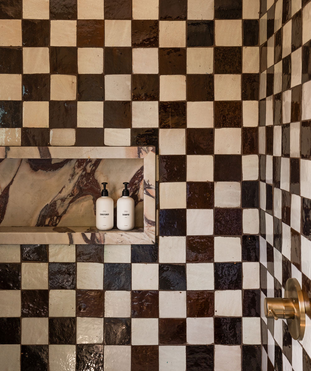 A shower wall covered in glossy brown and cream checkerboard tiles, featuring a built-in niche with two white pump bottles and a brass shower handle on the right.