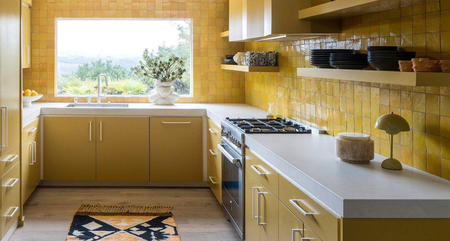 A modern kitchen with yellow cabinets and square yellow tiles, white countertops, a gas stove, open shelving with dark dishes, a window with a greenery view, and a small rug on the wooden floor.