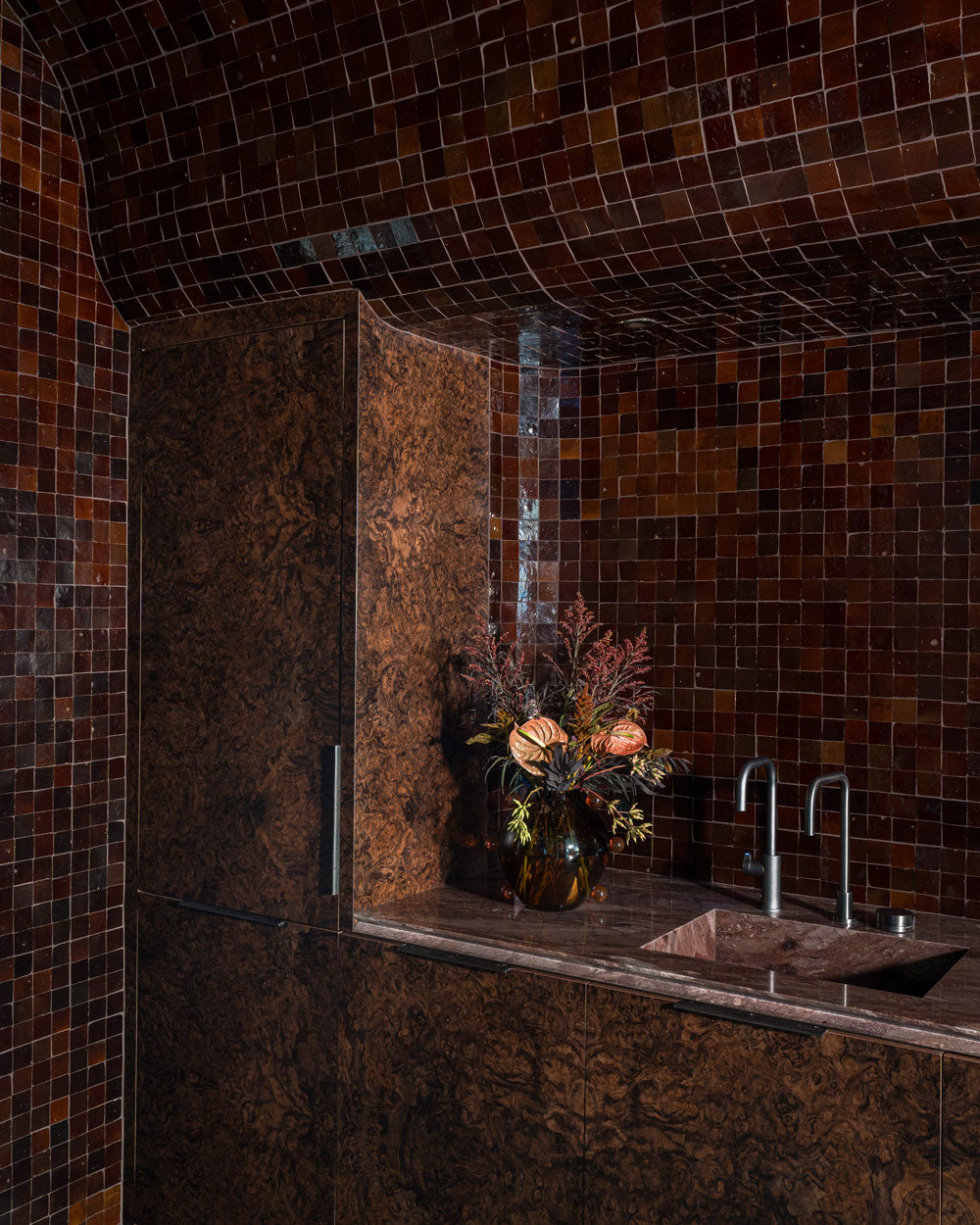 A kitchen corner with dark brown, glossy mosaic tiles on the walls and ceiling, a matching wood-textured cabinet, a double sink, and a vase with pink and green flowers on the countertop.