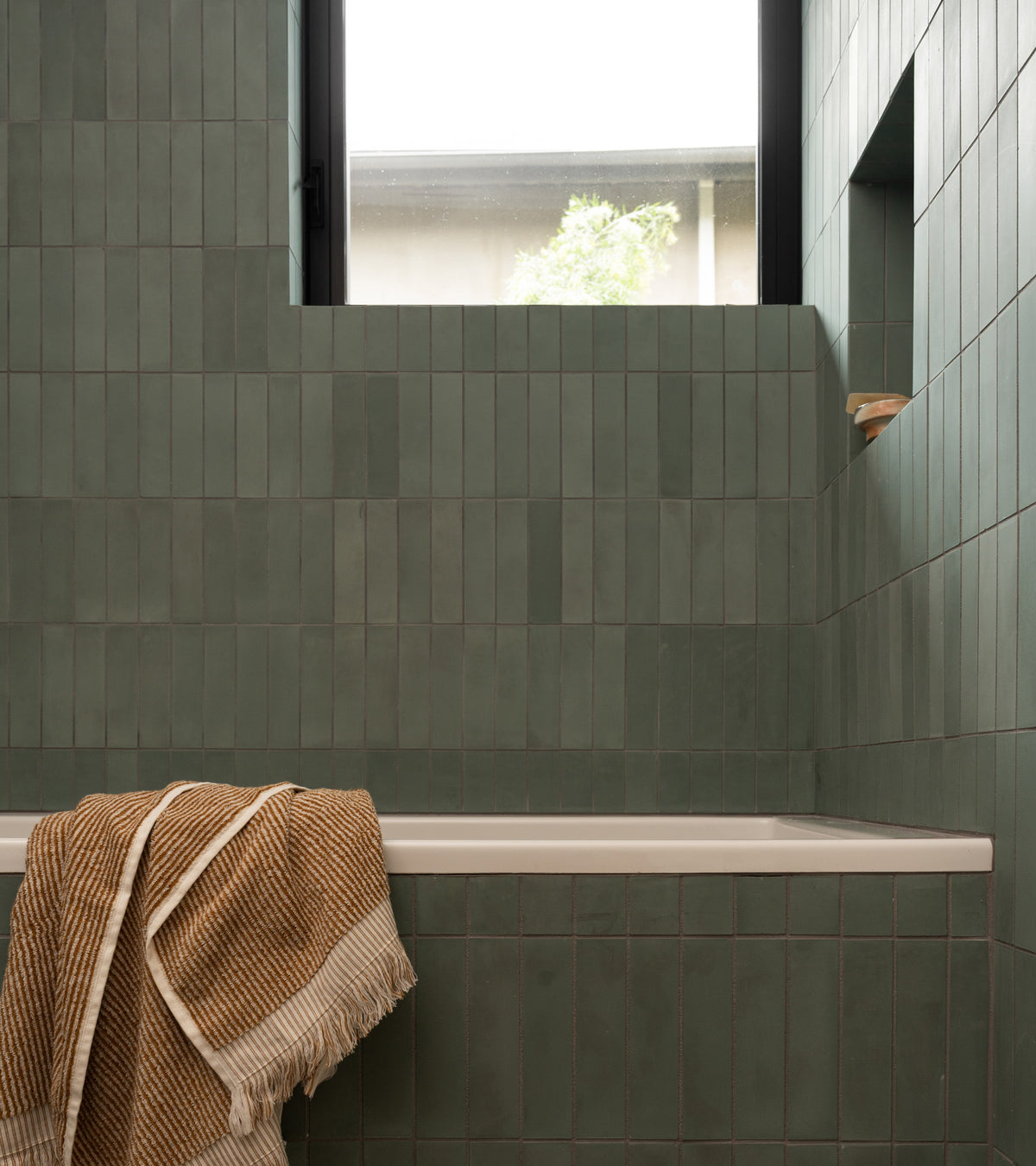 A bathroom features Zeppelin 2x8 green subway tiles by vendor-unknown on the walls, a white bathtub with a brown and white striped towel, a recessed shelf with soap, and a window above bringing in natural light.