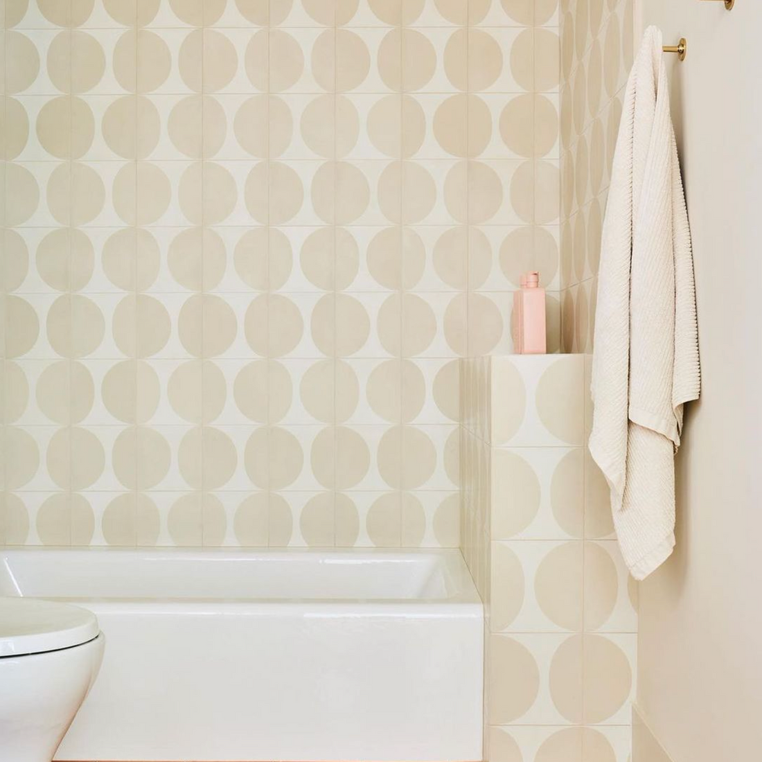 A modern bathroom features Pomelo Bone 8x8 beige handmade tile walls, a white bathtub, a peach floor, a pink bottle, and a white towel hanging on a hook.