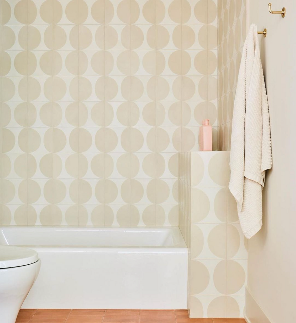 A modern bathroom features Pomelo Bone 8x8 beige handmade tile walls, a white bathtub, a peach floor, a pink bottle, and a white towel hanging on a hook.